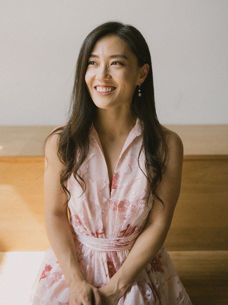 A smiling woman with long dark hair wearing a light pink floral dress is sitting on a wooden bench in soft natural light.